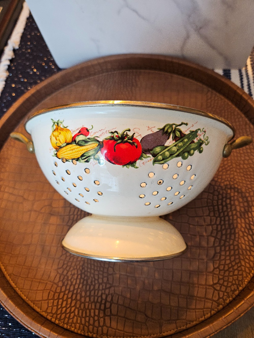 Vintage enamel colander features a vibrant fruit and vegetable design and ornate brass handles.